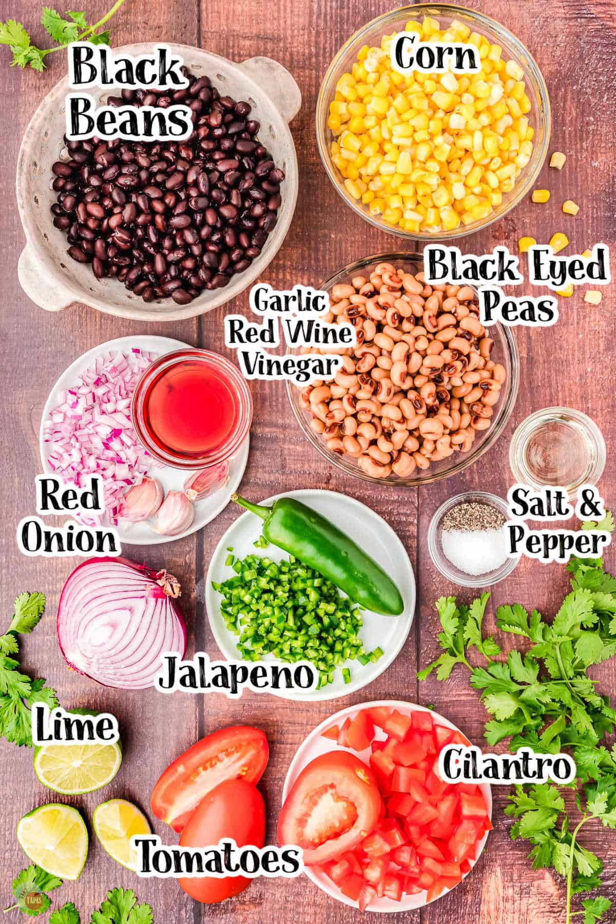 Wooden table with ingredients for Cowboy Caviar on it and labels denoting each one including  cilantri, tomatoes, limes, jalapeno, red onion, black eyed peas, garlic red wine vinegar, corn and black beans. 