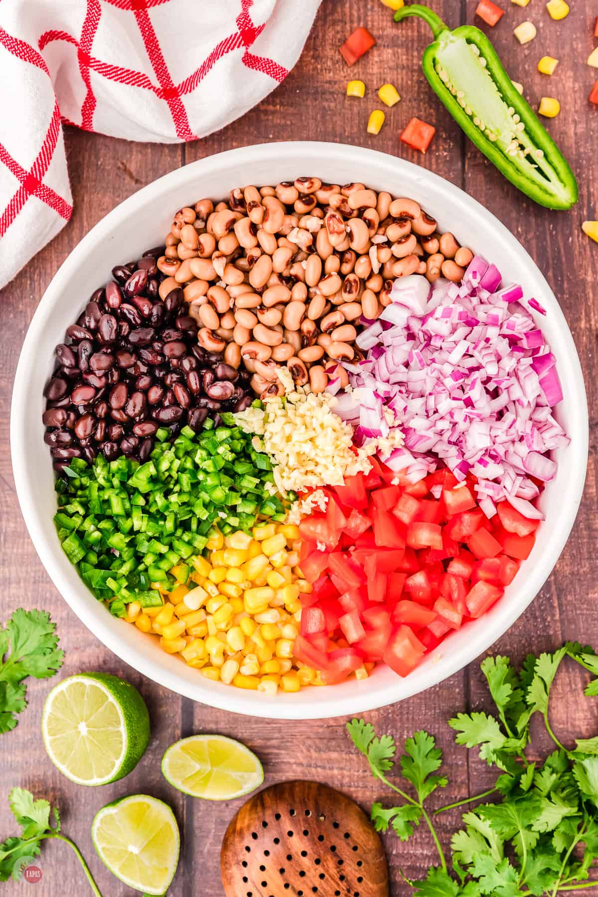 Top view of large white bowl on a table with all the chopped ingredients of cowboy caviar in it in sections, with some limes, cilantro and a jalapeno on the table next to it. 