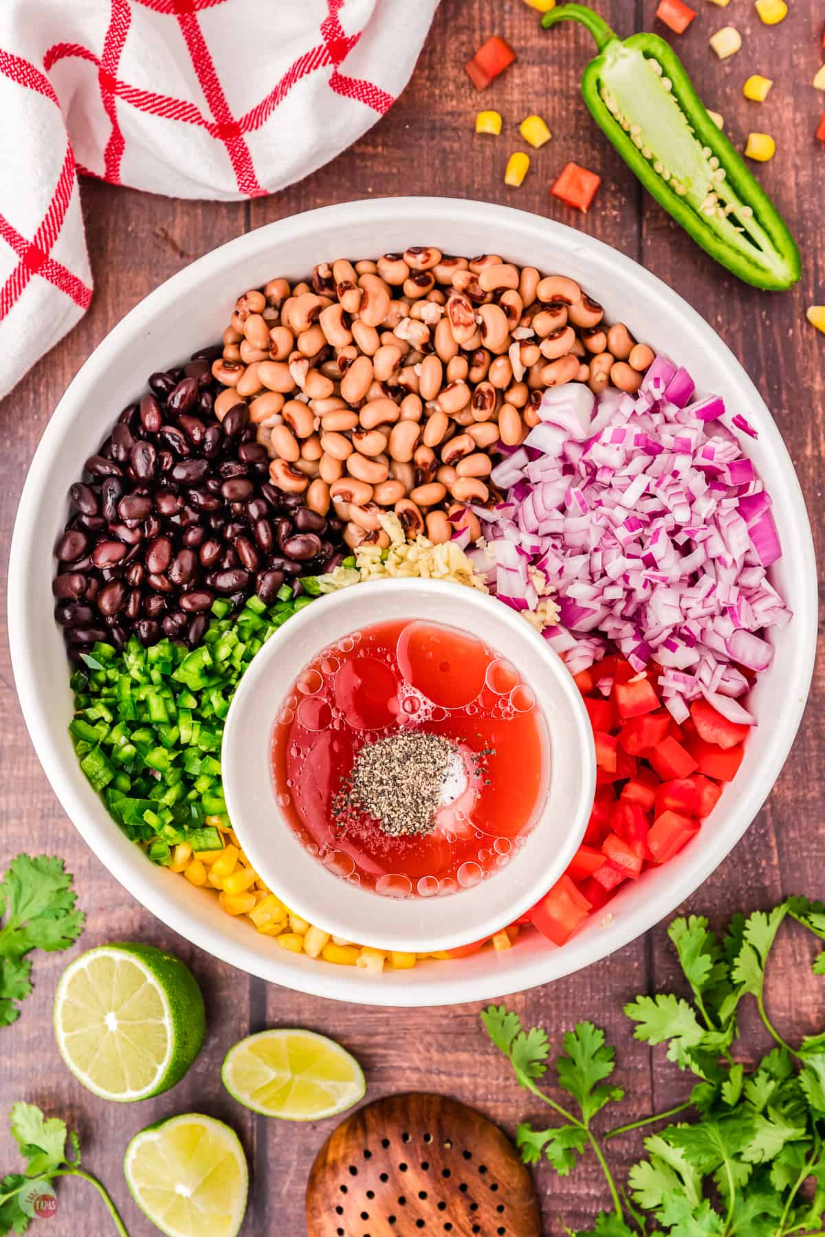 Top view of large white bowl on a table with all the chopped ingredients of cowboy caviar in it in sections, with a white ramekin sitting on top of the corn with the dressing ingredients in it. There are also some limes, cilantro and a jalapeno on the table next to it. 