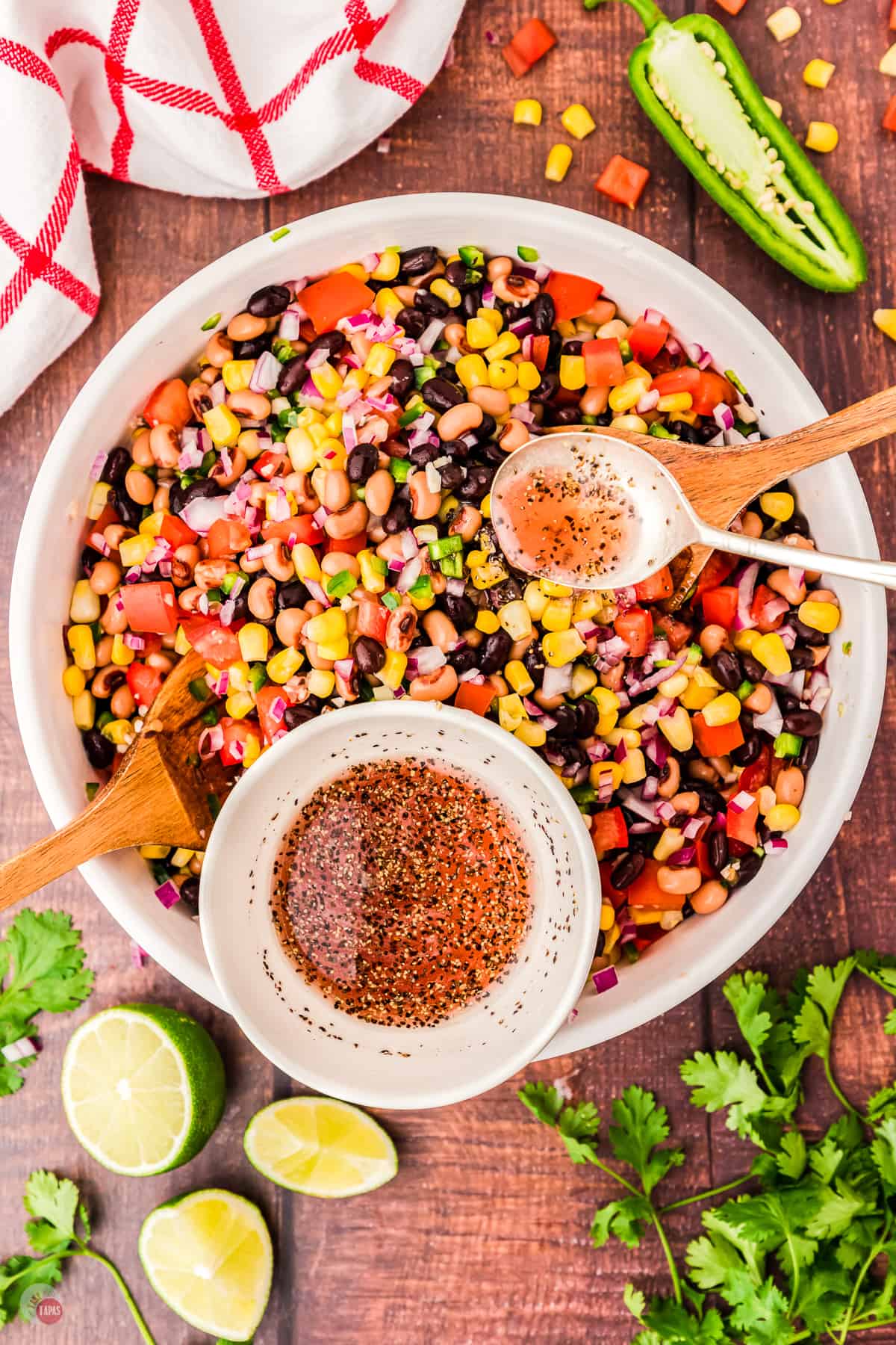 Top view of large white bowl on a table with all the chopped ingredients of cowboy caviar mixed up, with a white ramekin sitting on top of it with the dressing in it. There are also some limes, cilantro and a jalapeno on the table next to the bowl. 