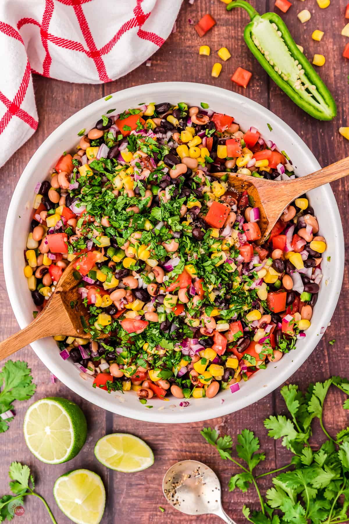 Top view of a large white bowl filled with cowboy caviar salad with two wooden serving utensils in it on either side. There are also some limes, cilantro and a jalapeno on the table next to the bowl. 