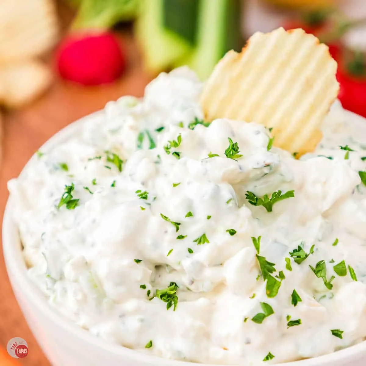 A small white bowl on a worktop filled with green onion dip garnished with parsley with a chip being lifted in mid-air that has some dip on it.