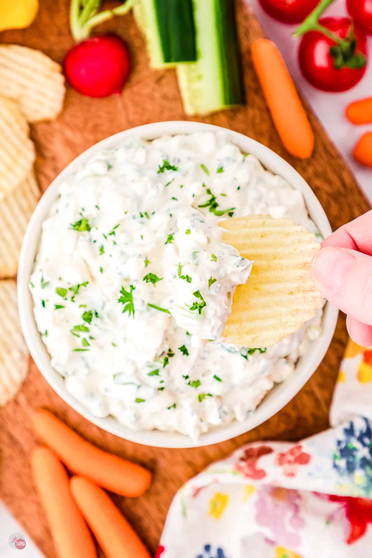 Top view of a small white bowl on a worktop filled with green onion dip garnished with parsley with a chip being lifted in mid-air that has some dip on it.