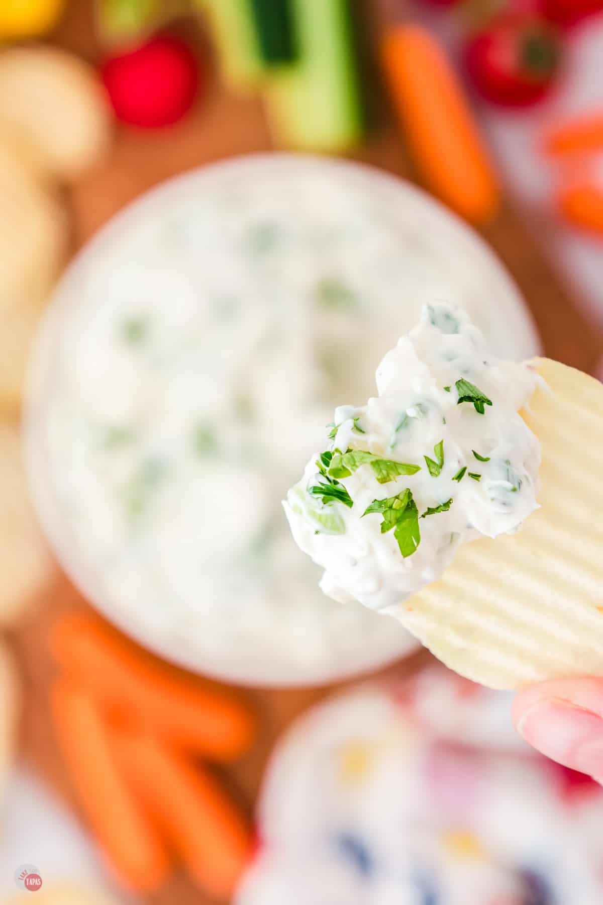 Top view of a small white bowl blurred in the background on a worktop filled with green onion dip garnished with parsley with a chip in focus being lifted in mid-air that has some dip on it.
