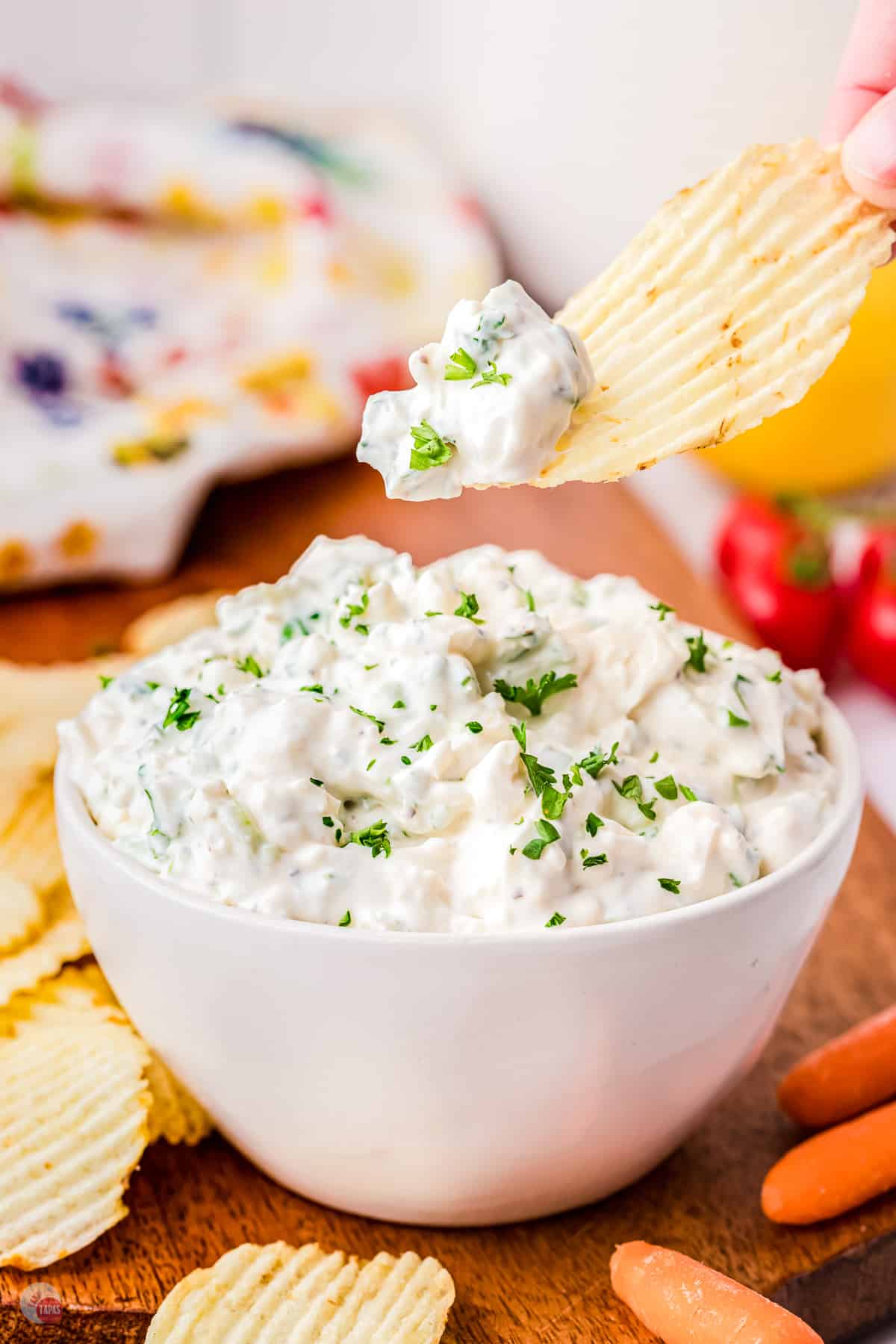 A small white bowl on a worktop filled with green onion dip garnished with parsley with a chip being lifted in mid-air that has some dip on it. 