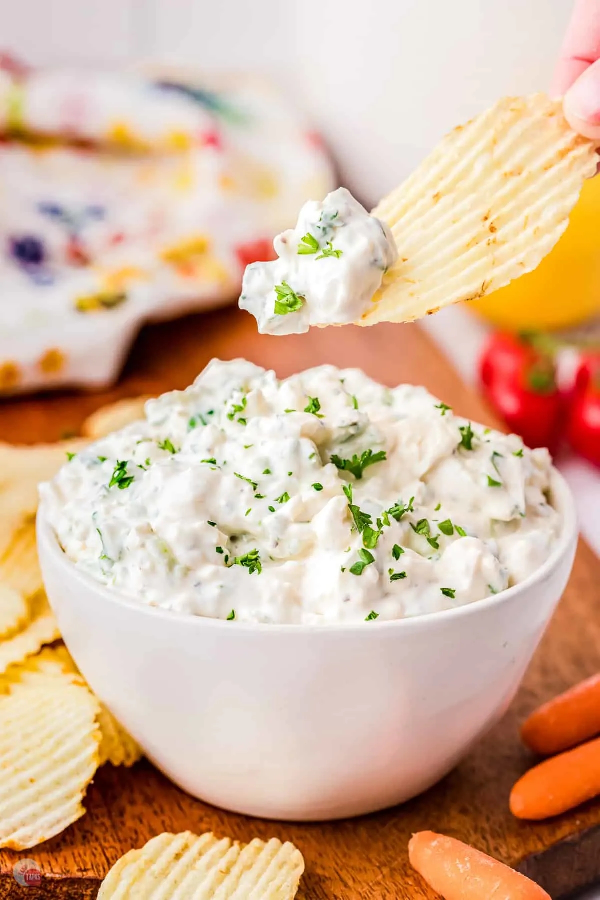 A small white bowl on a worktop filled with green onion dip garnished with parsley with a chip being lifted in mid-air that has some dip on it.