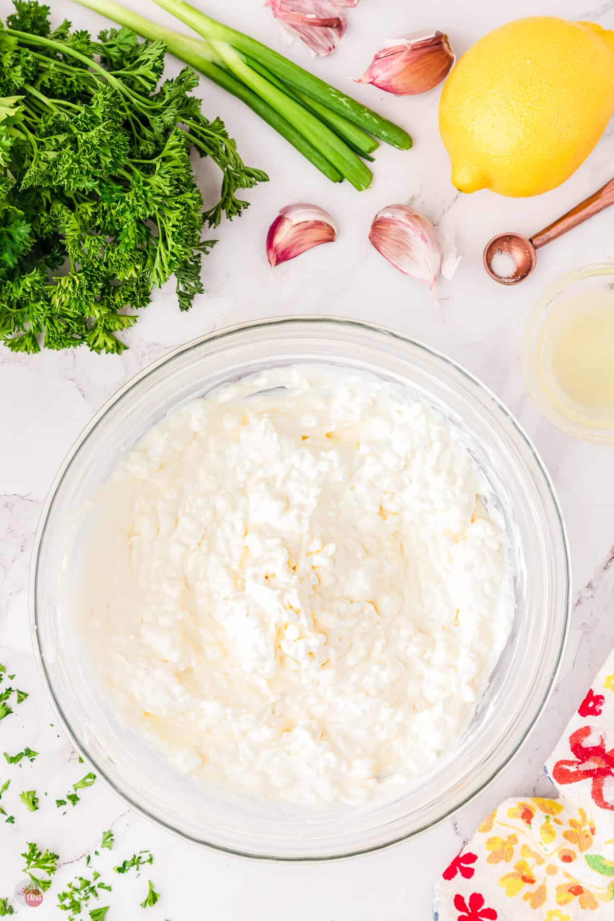 Top view of a large glass bowl on a worktop filled with cream cheese and sour cream mixture, surrounded by lemon, green onion, and fresh parsley. 