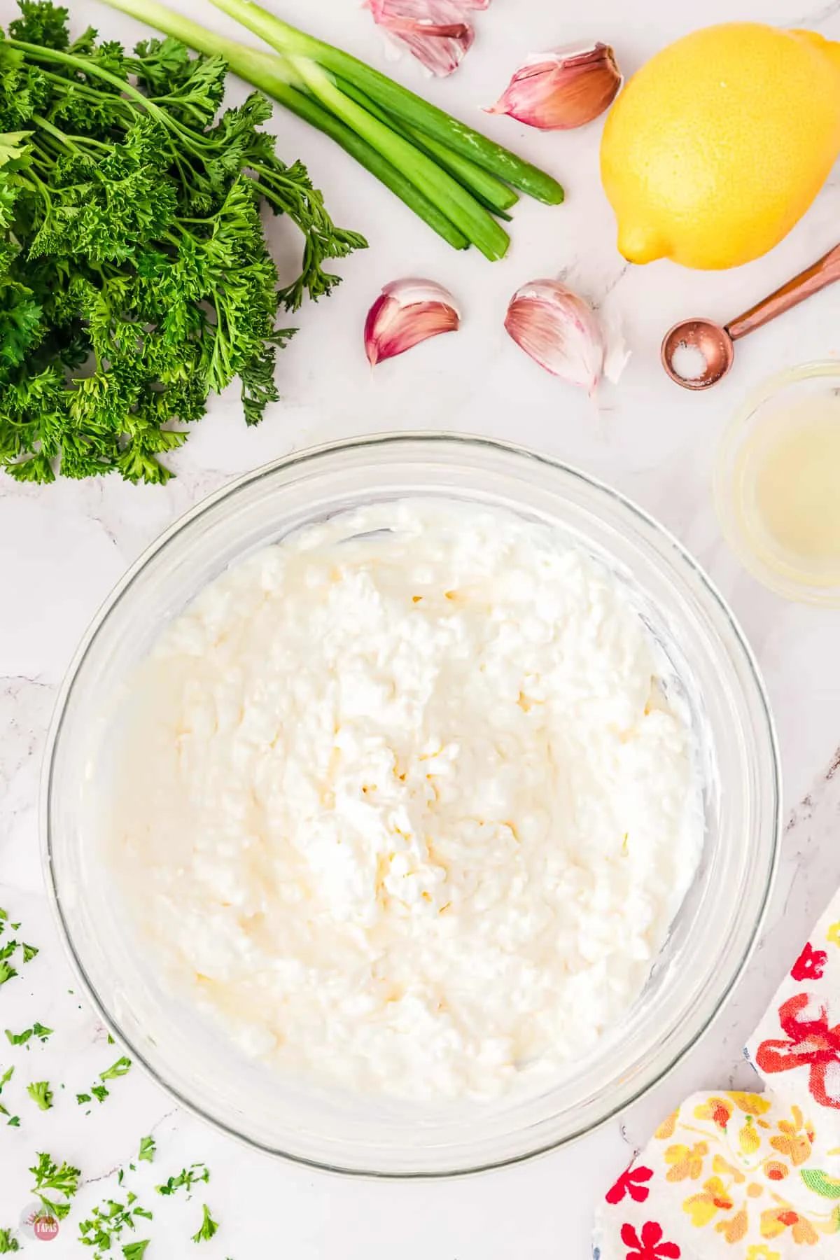 Top view of a large glass bowl on a worktop filled with cream cheese and sour cream mixture, surrounded by lemon, green onion, and fresh parsley.