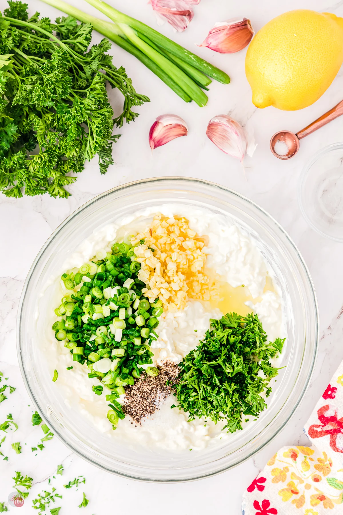 Top view of a large glass bowl on a worktop filled with unmixed green onion dip ingredients, surrounded by lemon, green onion, and fresh parsley. 