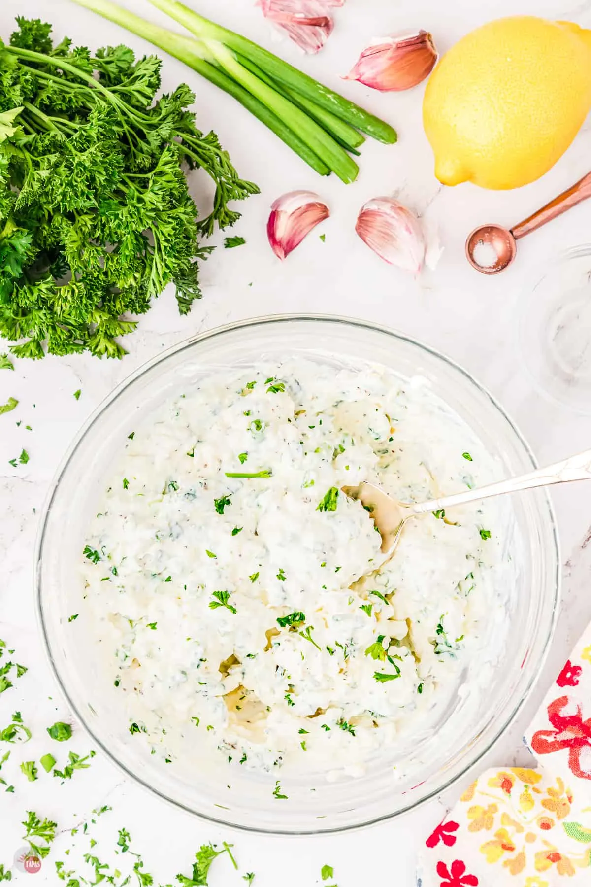 Top view of a large glass bowl on a worktop filled with green onion dip with a spoon in it surrounded by lemon, green onion, and fresh parsley.