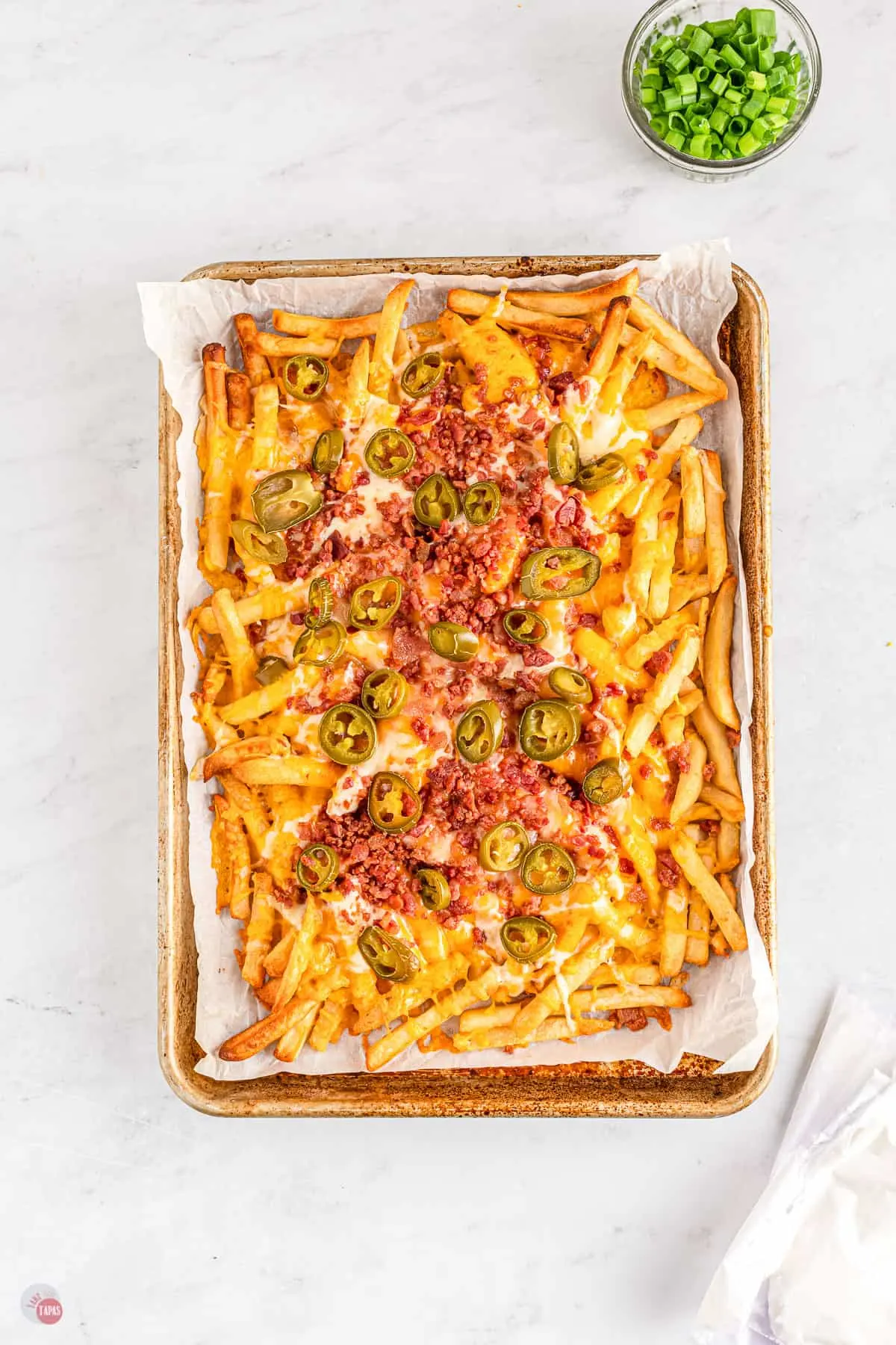 A baking tray lined with parchment paper filled with baked french fries topped with shredded cheese, bacon crumbles, and sliced jalapeños.