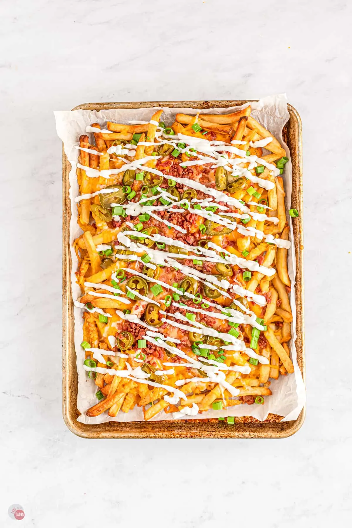 A baking tray lined with parchment paper filled with baked french fries topped with shredded cheese, bacon crumbles, sliced jalapeños, chopped green onion, and drizzled in sour cream.