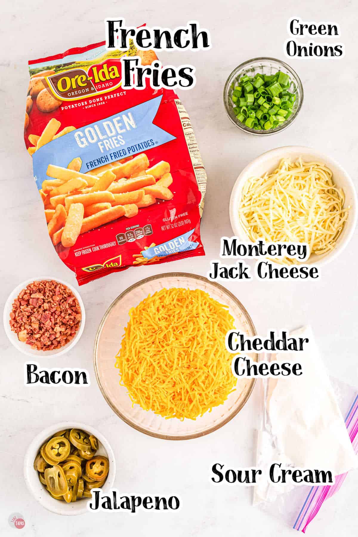 Top view of ingredients for loaded fries including a bag of frozen fries, and then bowls in various sizes filled with green onions, Monterey Jack cheese, bacon crumbles, jalapeños and at the bottom of the picture there is a freezer bag with sour cream in it.