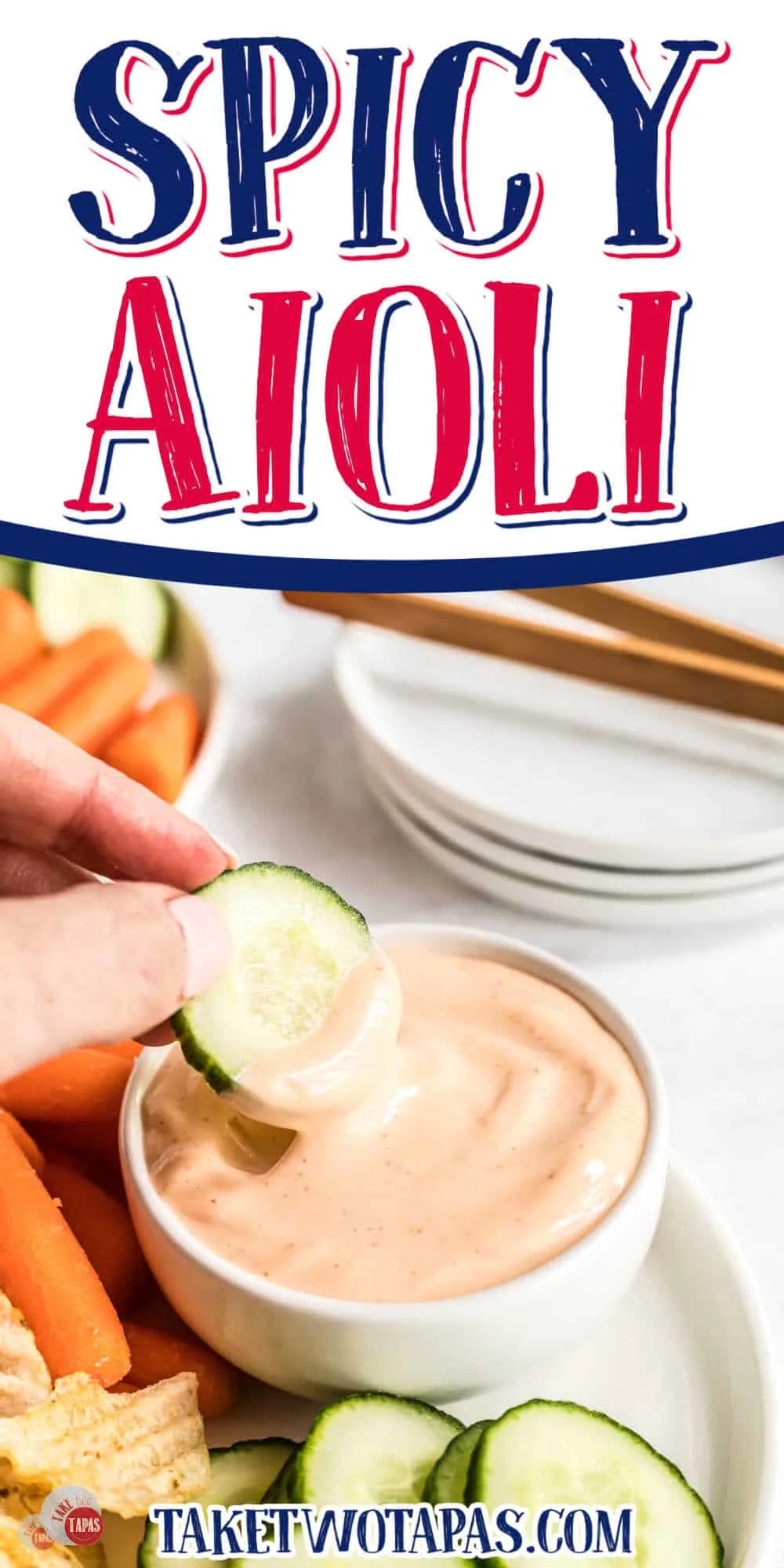 Spicy aioli sauce in small bowl on a white plate with fresh baby carrots and slices of cucumber next to it, and there is a hand dipping a slice of cucumber into the sauce and lifting it out mid-air. There is text on the top part of the image that reads "Spicy Aioli".