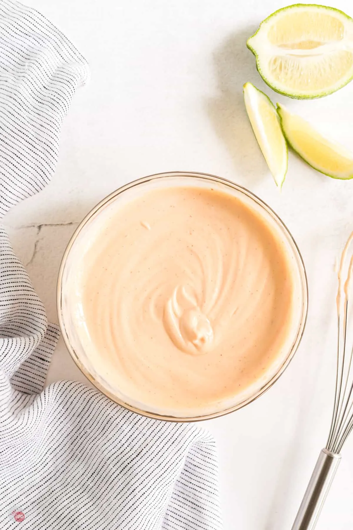 Spicy aioli in a small bowl on a worktop with half a lime and lime slices and a whisk next to it.