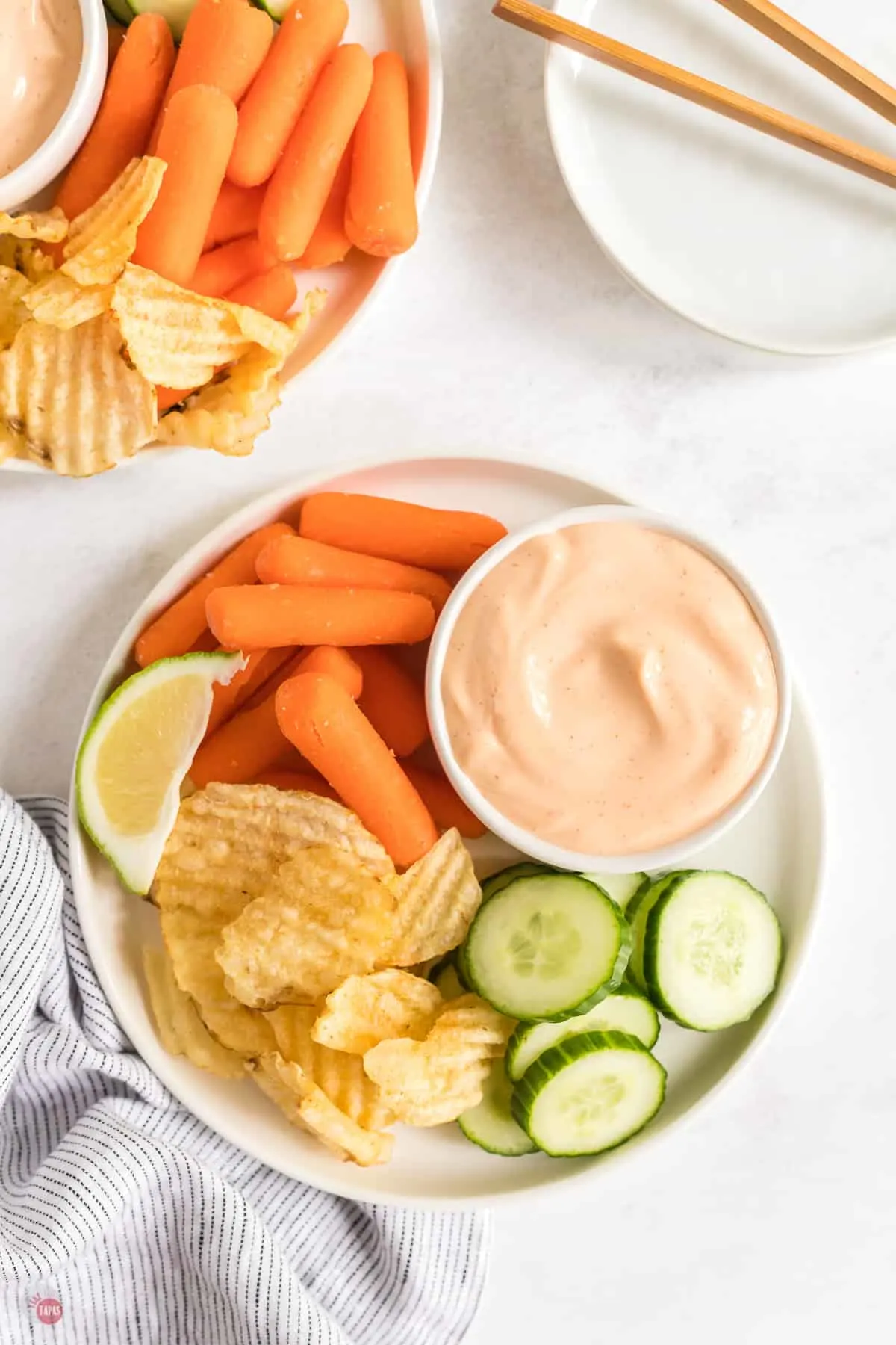 Top view of spicy aioli sauce in a small bowl on a white plate with fresh baby carrots and slices of cucumber and chips next to it.