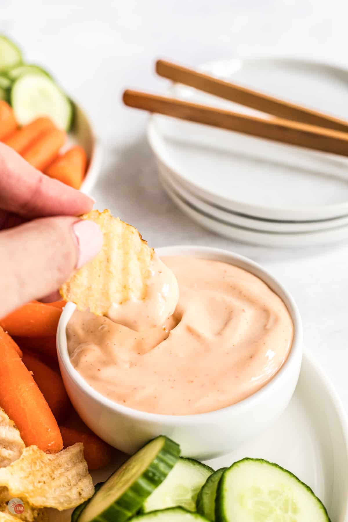 Spicy aioli sauce in small bowl on a white plate with fresh baby carrots and slices of cucumber next to it, and there is a hand dipping a crinkle chip into the sauce and lifting it out mid-air. 