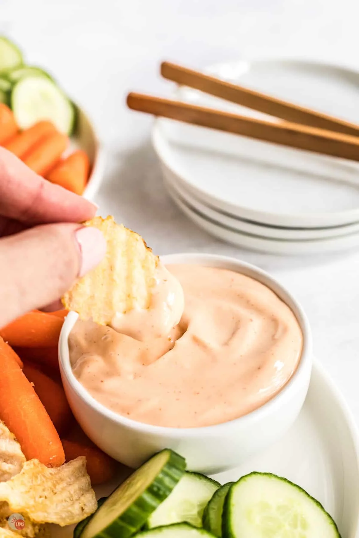 Spicy aioli sauce in small bowl on a white plate with fresh baby carrots and slices of cucumber next to it, and there is a hand dipping a crinkle chip into the sauce and lifting it out mid-air. 