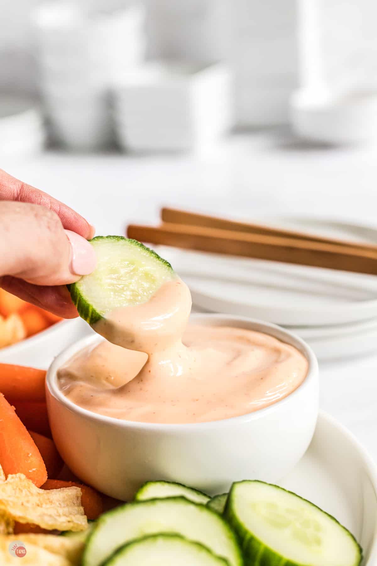 Spicy aioli sauce in small bowl on a white plate with fresh baby carrots and slices of cucumber next to it, and there is a hand dipping a slice of cucumber into the sauce and lifting it out mid-air. 