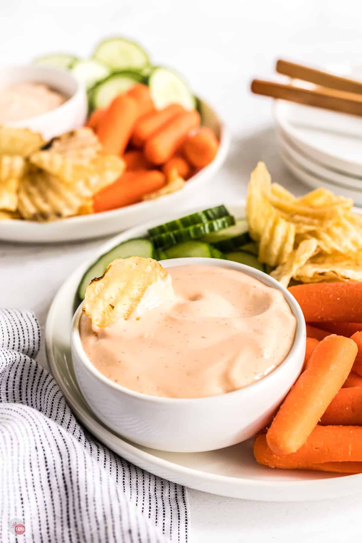 Spicy aioli sauce in small bowl with a chip in the sauce, on a white plate with fresh baby carrots, slices of cucumber, and other chips next to it. 