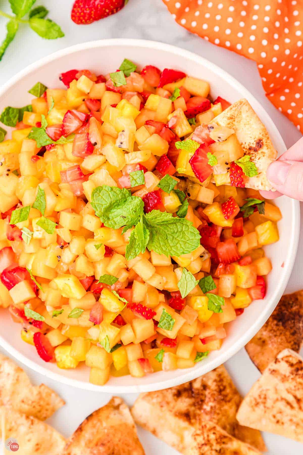 hand holding a chip in a bowl of fresh fruit salsa with mint leaves on top