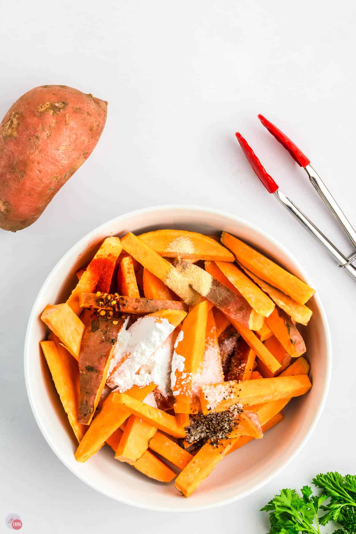 sweet potato wedges in a white bowl with cornstarch and seasonings on top. A pair of red tongs next to the bowl