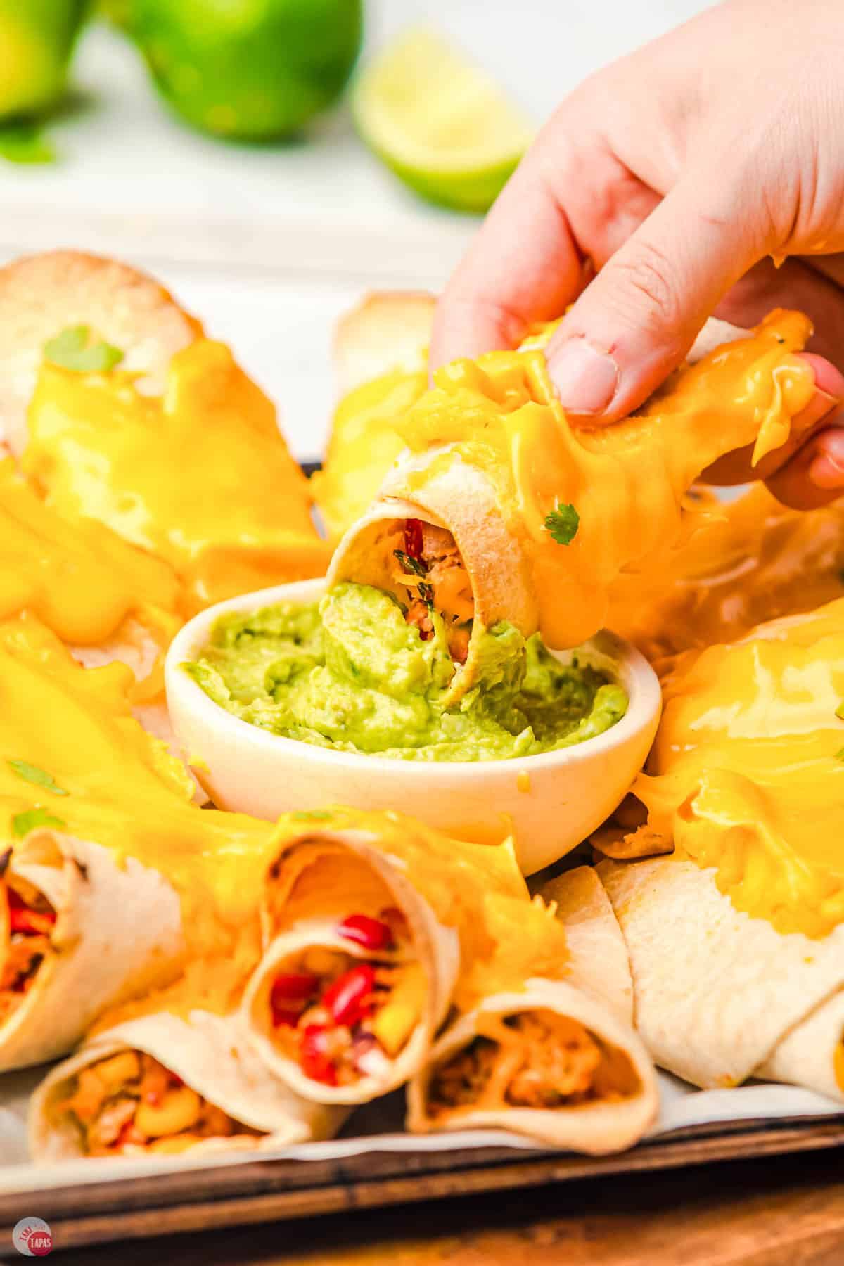 hand holding a rolled taco, covered in cheese, and dipping in some guacamole
