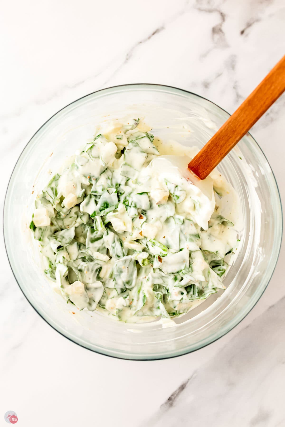 spinach dip being mixed in a clear bowl with a spatula