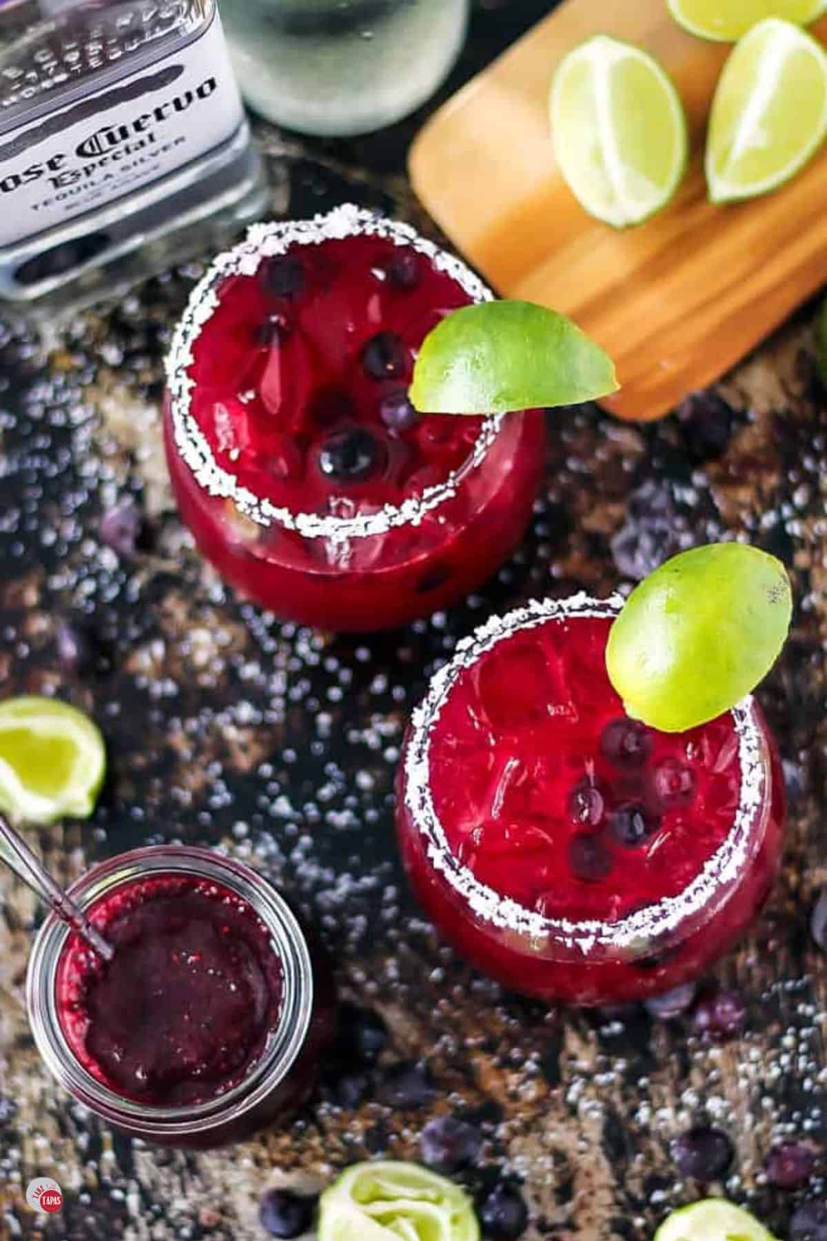 two glasses of blueberry margaritas and a cutting board with limes