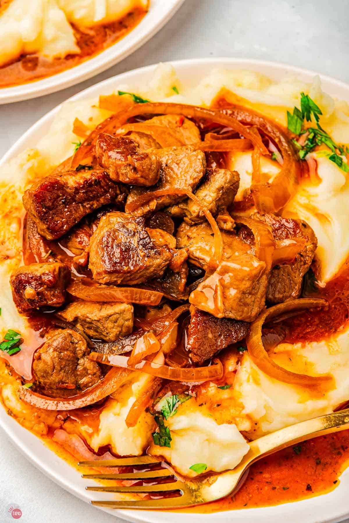 plate of mashed potatoes with pieces of steak, onions, and gravy on top