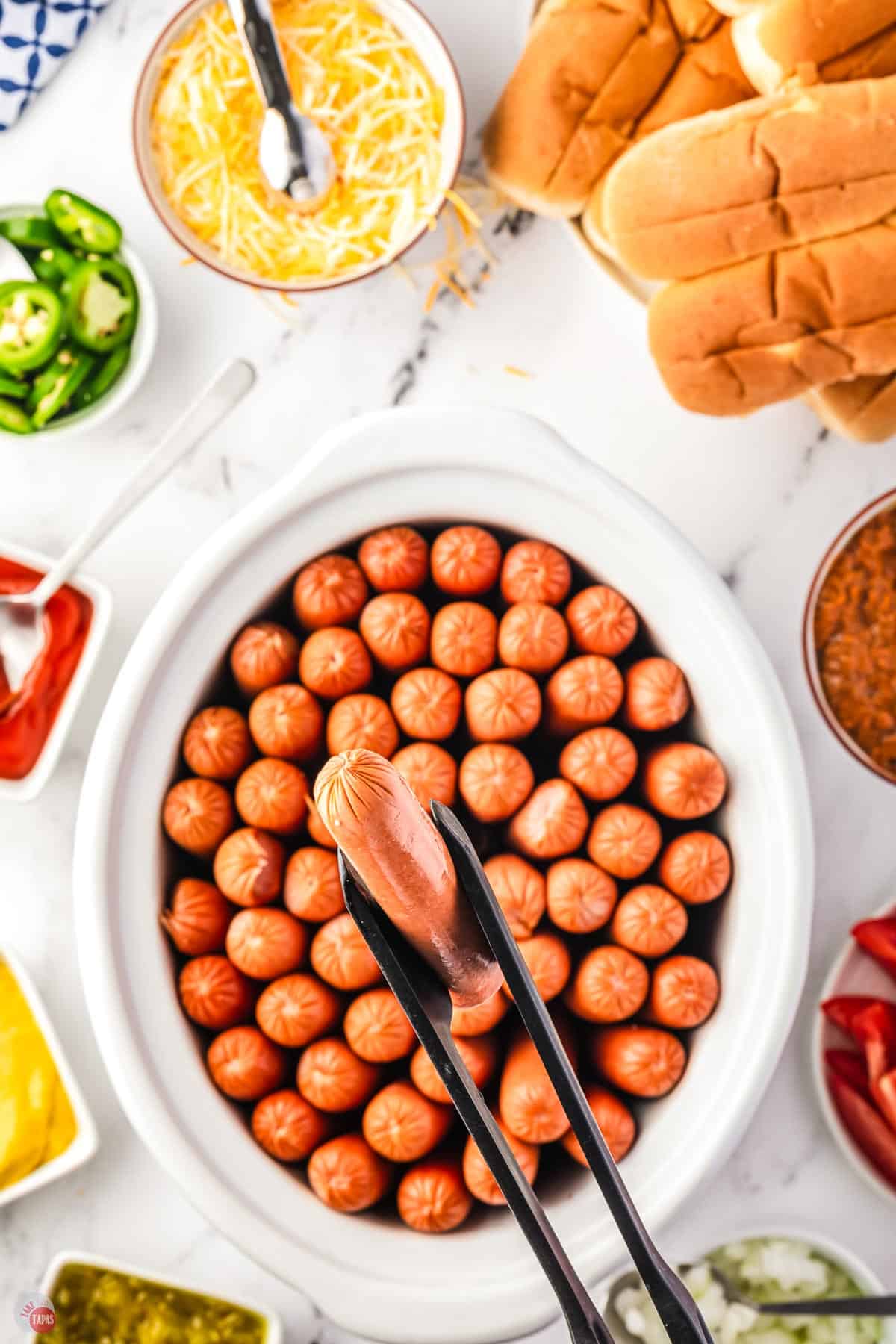 close up of tongs holding a single hot dog over a bowl of extra hot dogs