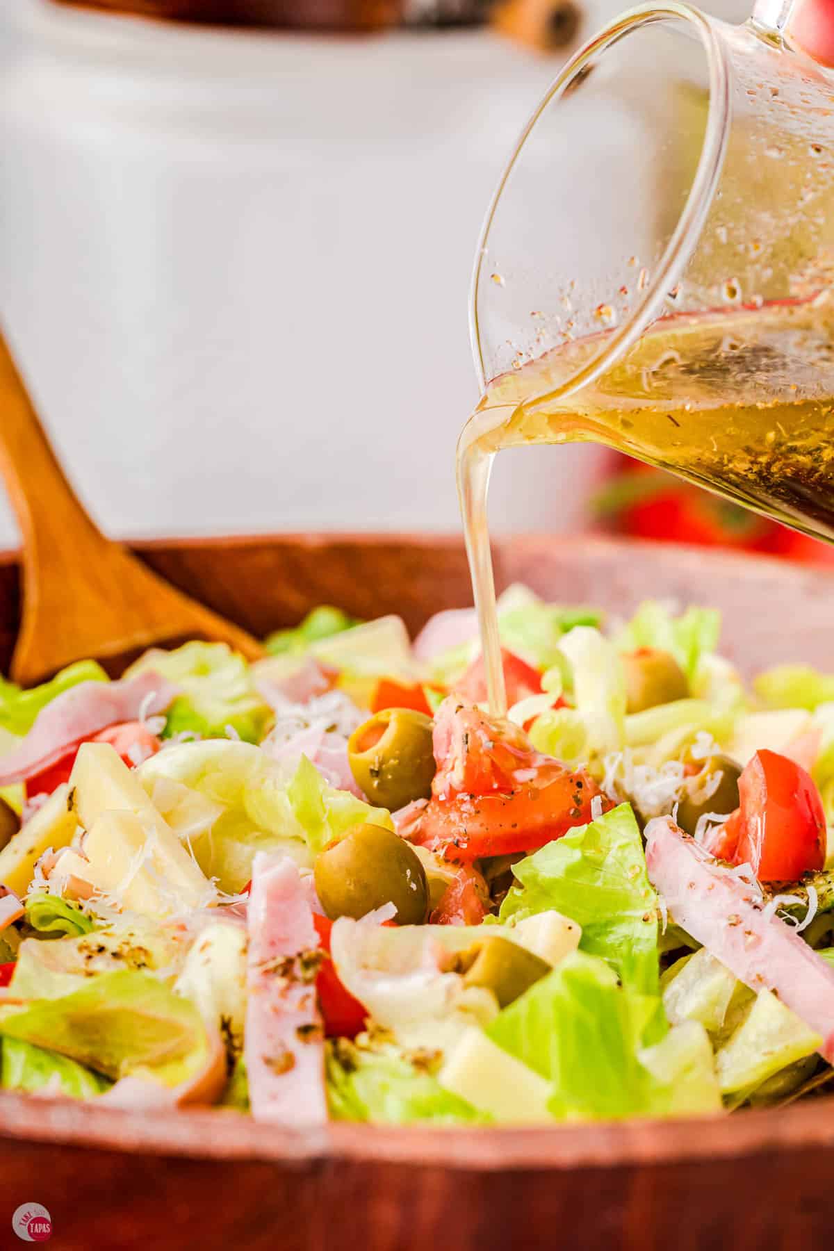 A hand is pouring dressing over a salad in a wooden bowl.
