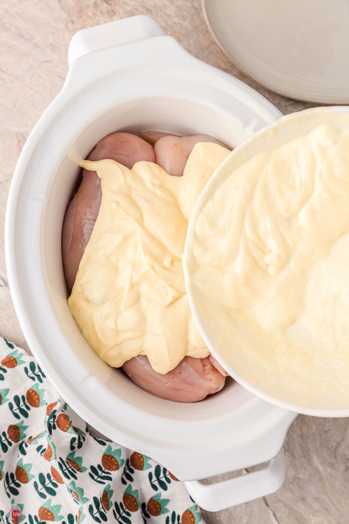 white crockpot with chicken in the bottom and a sauce being poured over the top