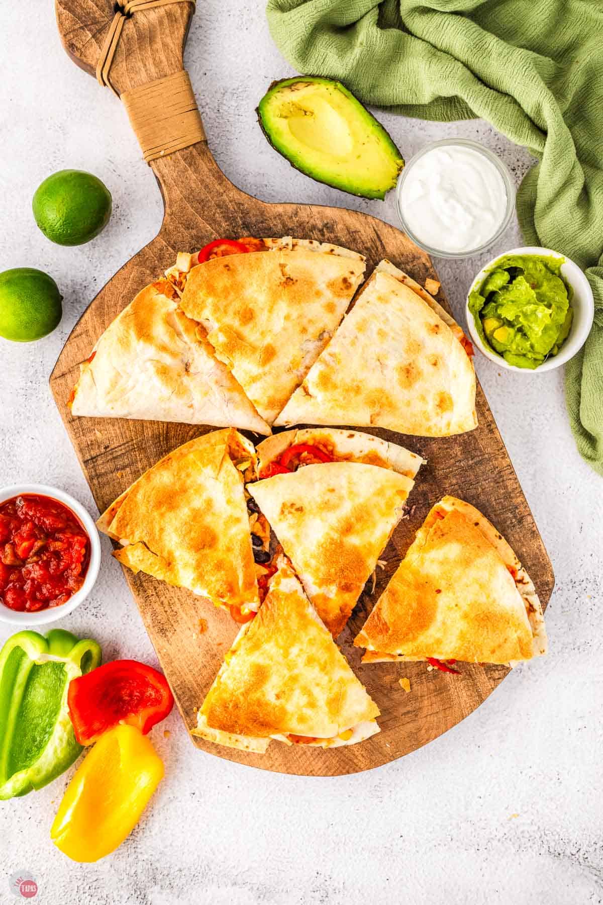 wood board with wedges of chicken quesadilla on it with bowls of toppings