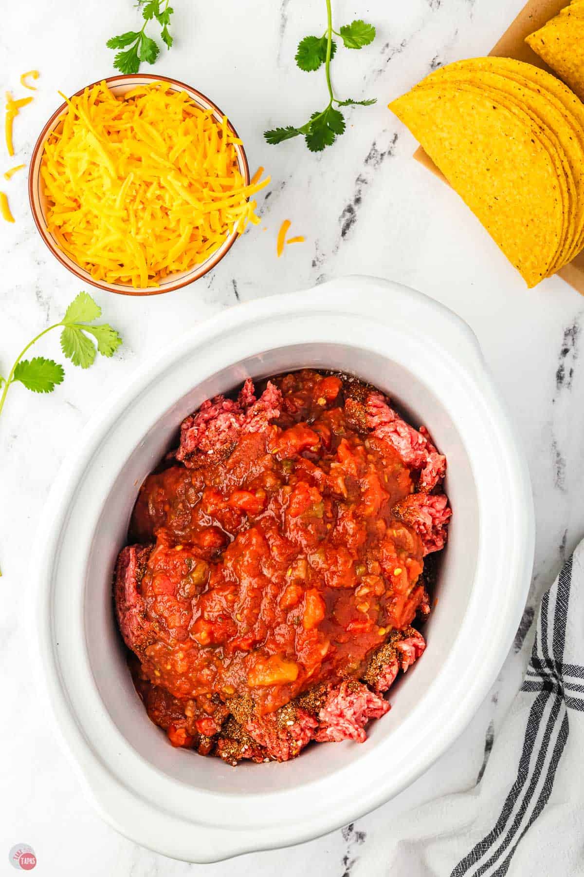 salsa on top of uncooked ground beef in a white crock pot bowl with taco shells next to it