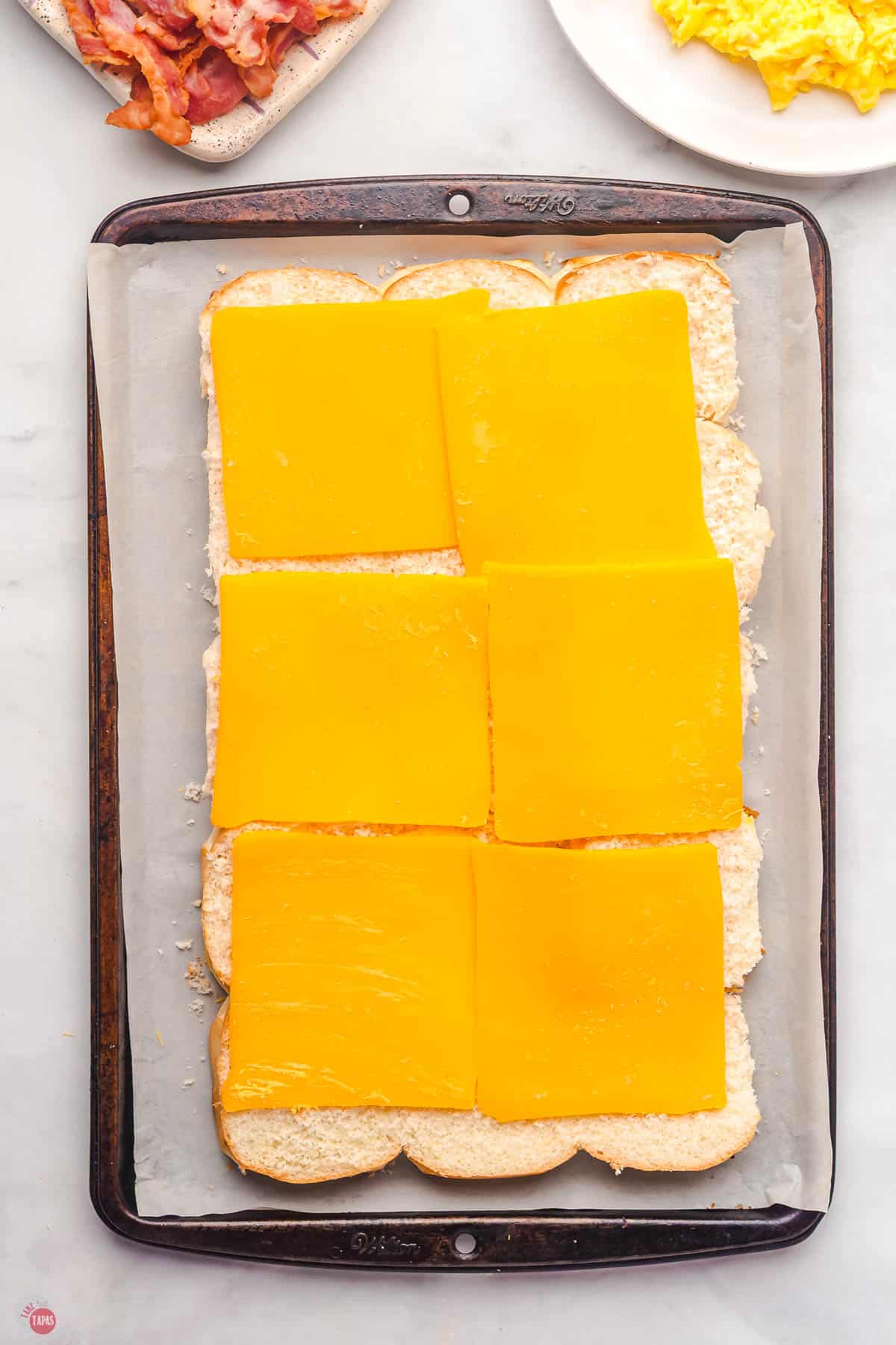 slices of cheese on top of slider buns on a baking sheet