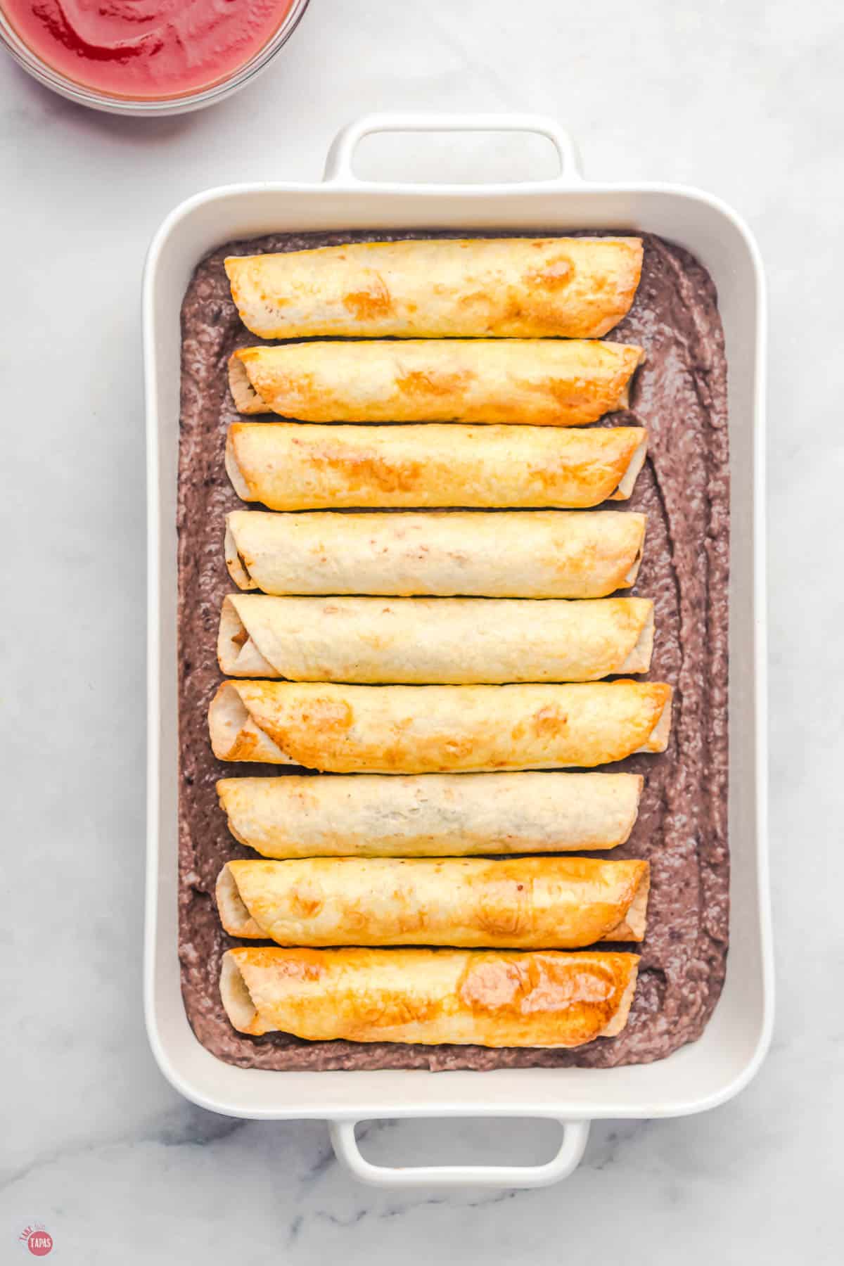refried beans in a baking dish with a layer of taquitos on top