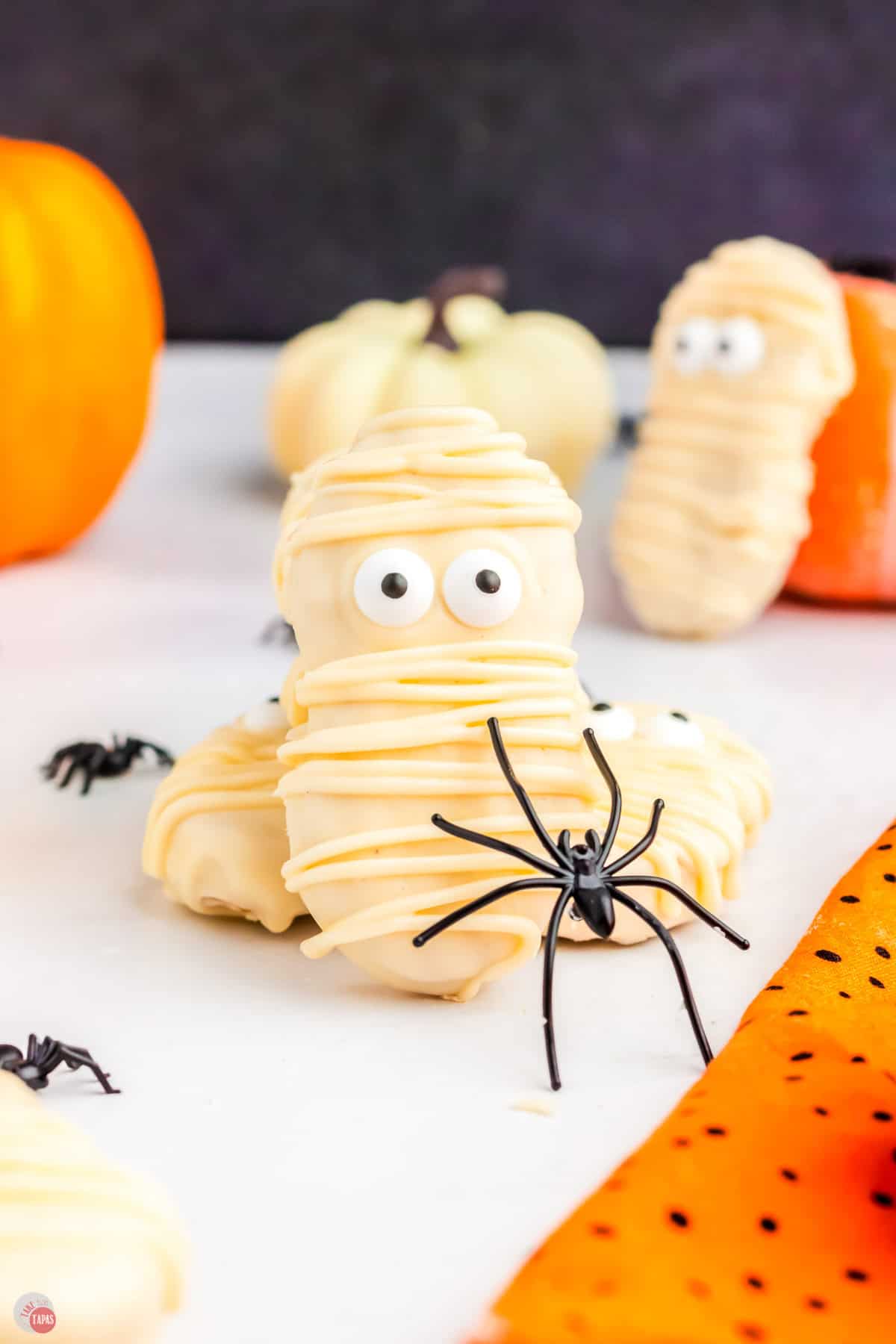 two mummy cookies on a white counter with pumpkins and a spider