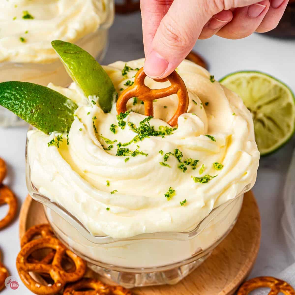 clear bowl overflowing with dip topped with lime zest and a salted pretzel on the top