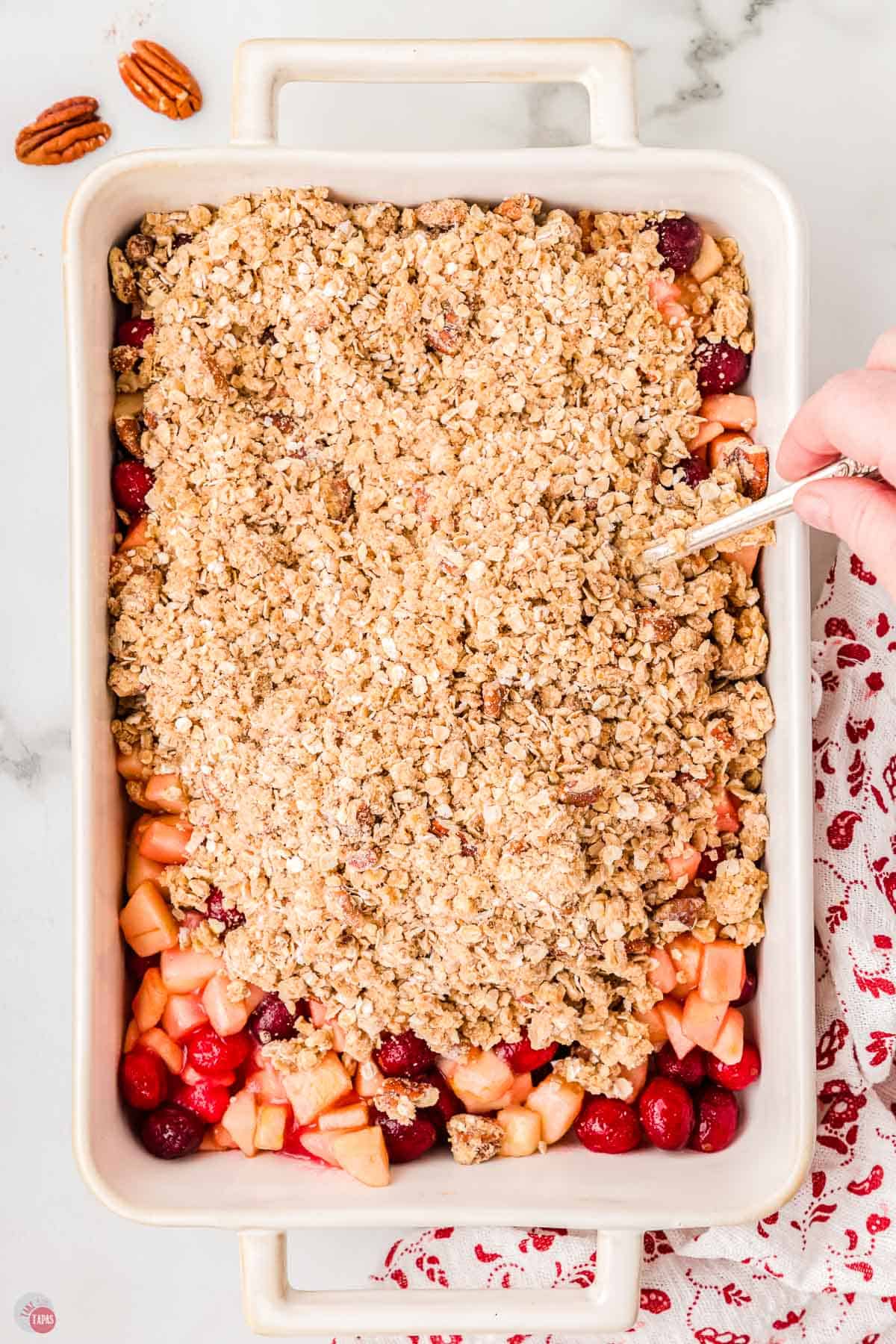 casserole dish with a hand spreading oatmeal over the top of apples and cranberries