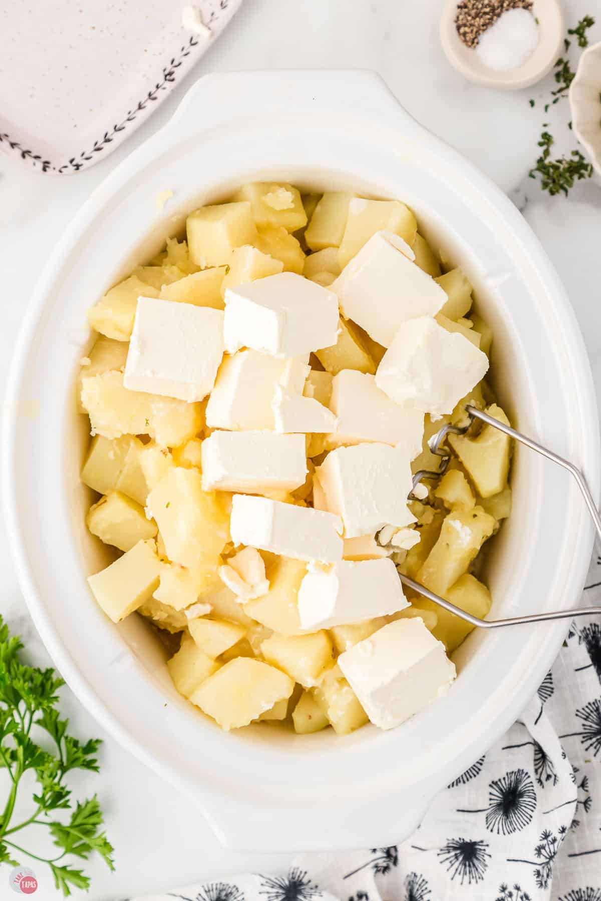 white slow cooker filled with diced potatoes and cubes of cream cheese on top