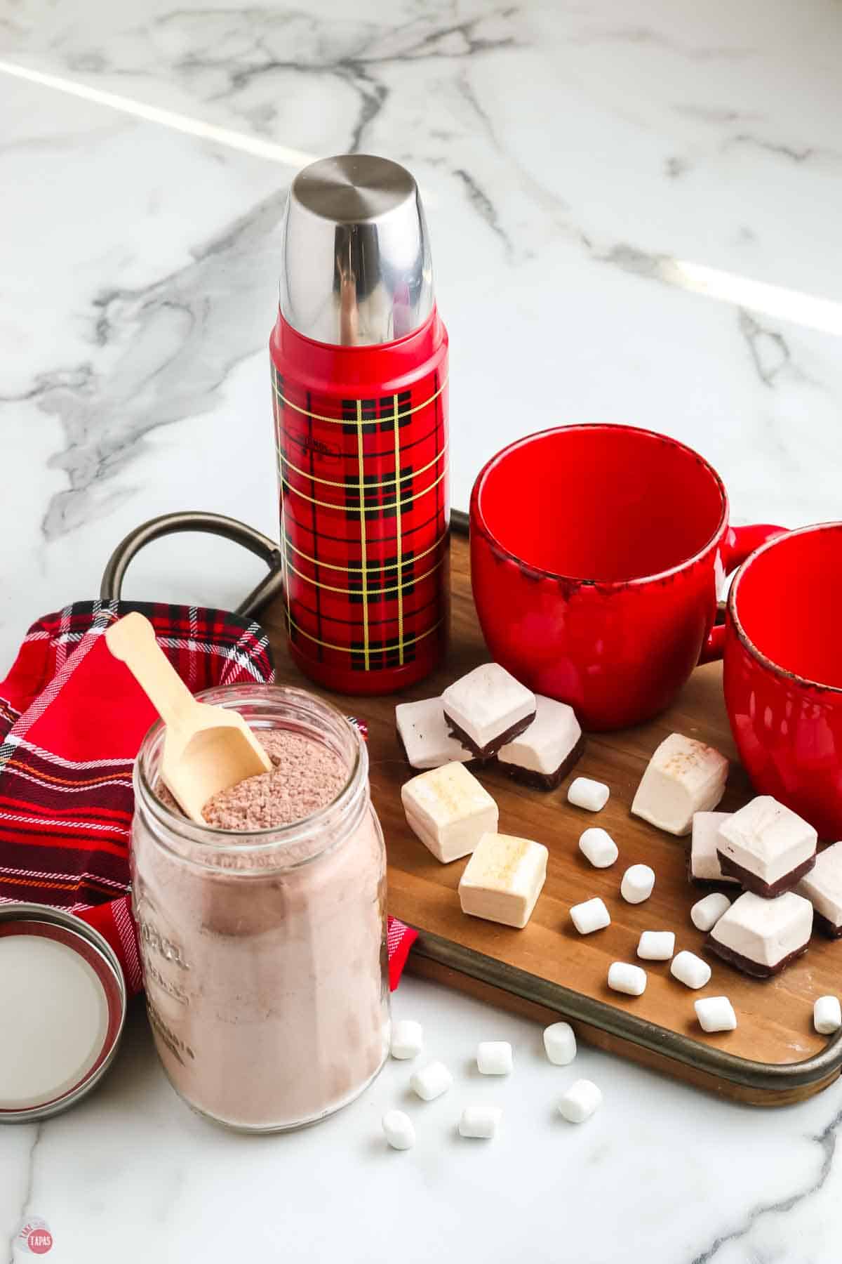 board with cocoa mix and marshmallows with cups and a thermos