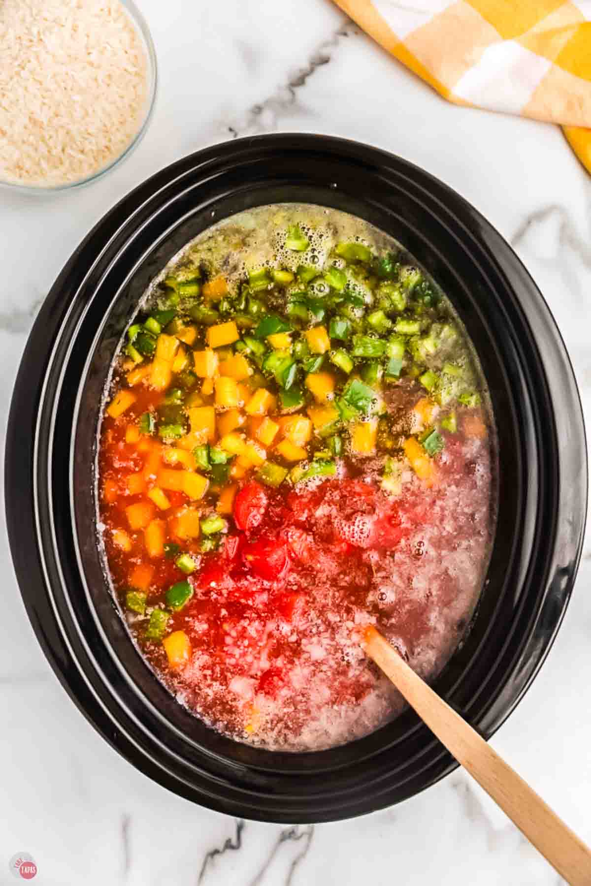 crock pot with bell peppers and tomatoes being stirred with a wood spoon