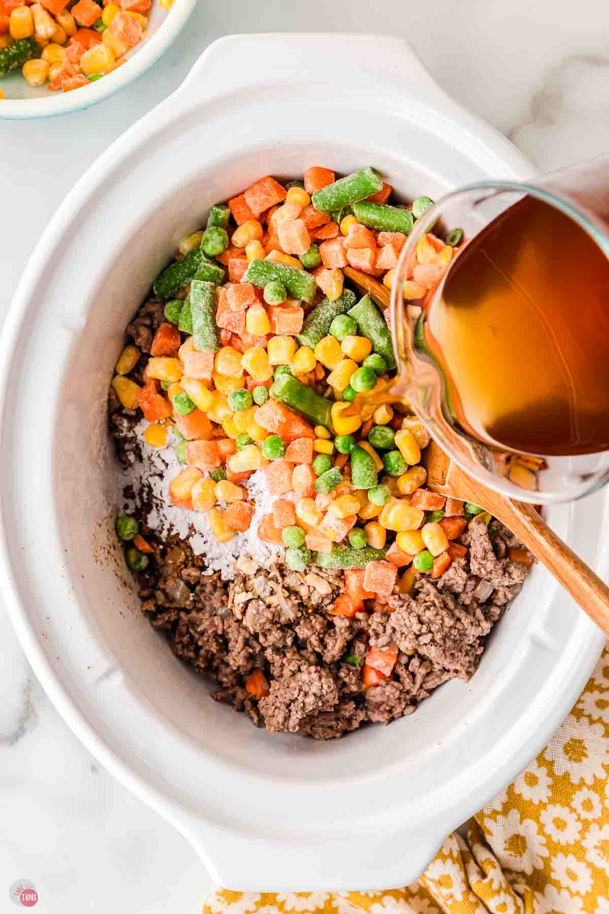white crock pot with ground beef, onions, and mixed vegetables in it and broth being poured over it