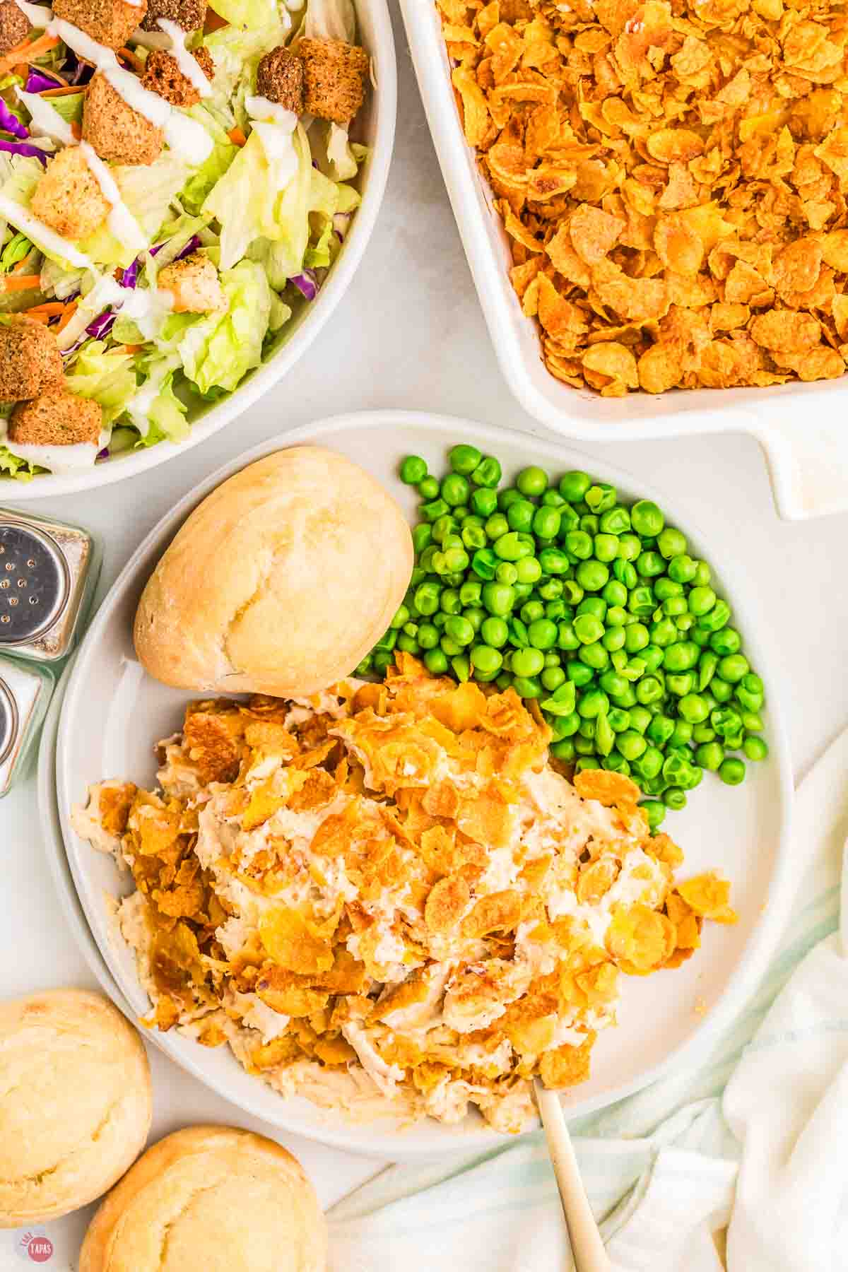 overhead picture of chicken casserole on a white plate with green peas and a dinner roll next to a bowl of salad