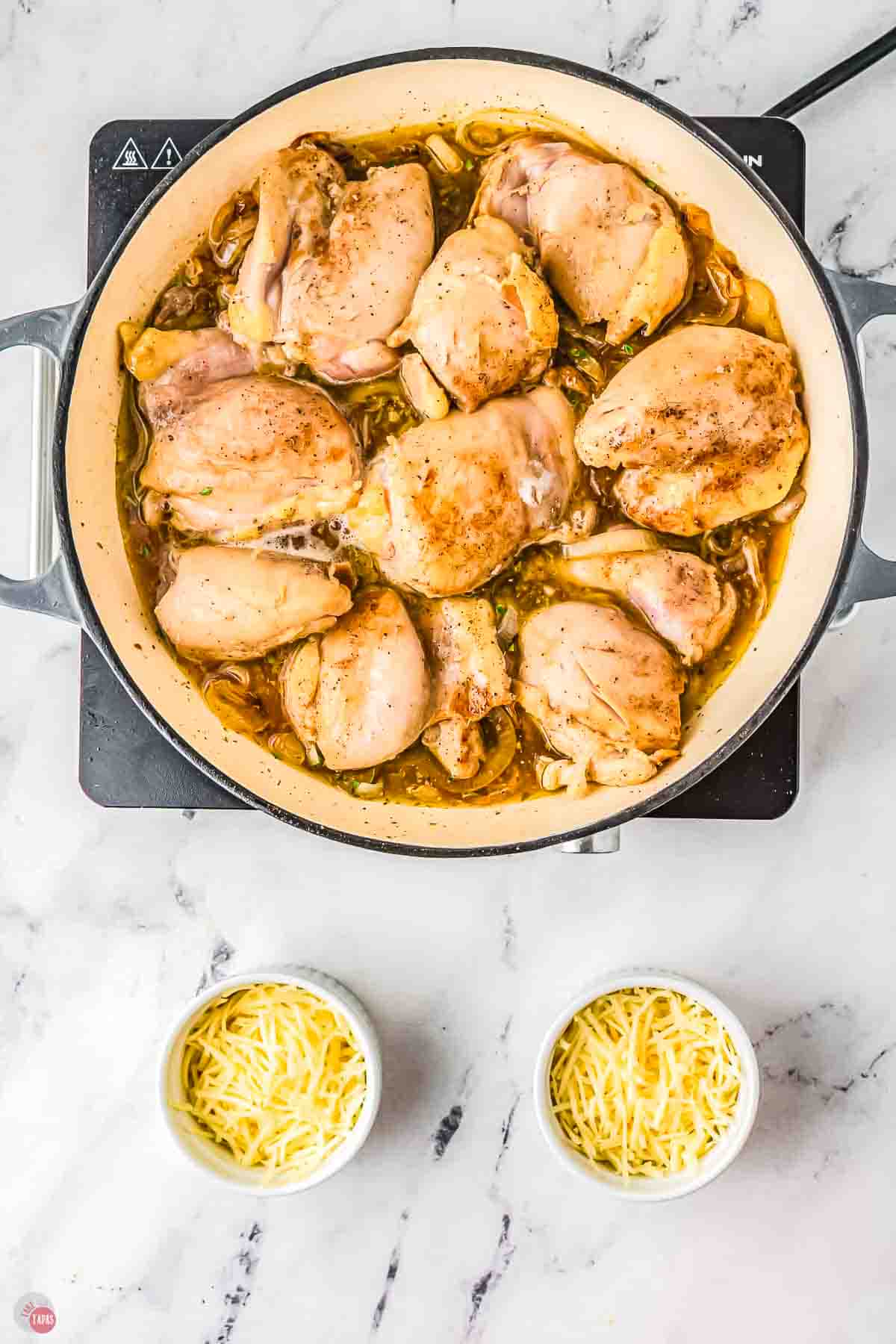 chicken thighs cooking in a white pan with two small bowls of cheese