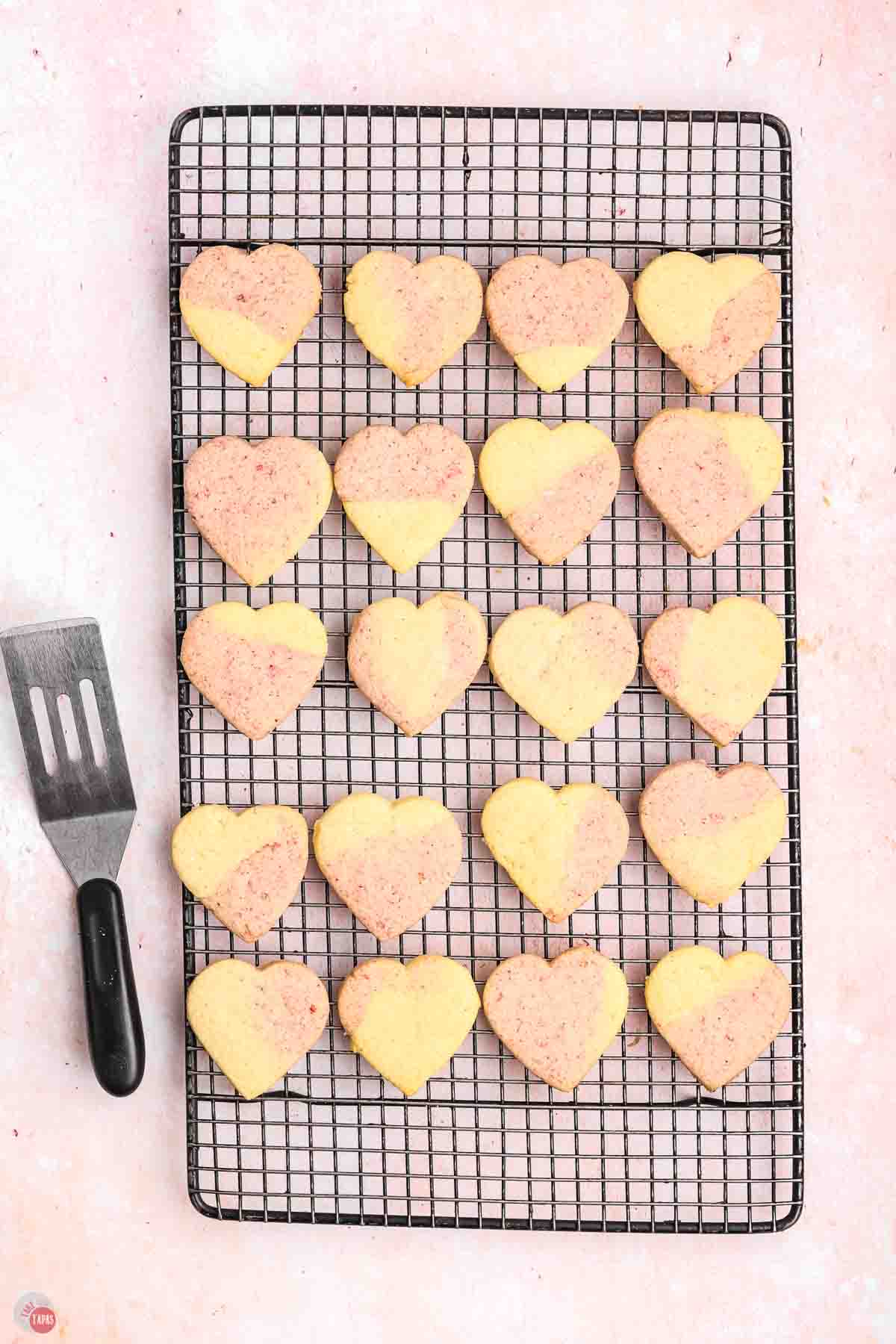 pink and yellow sugar cookies on a black wire cooling rack with a small spatula