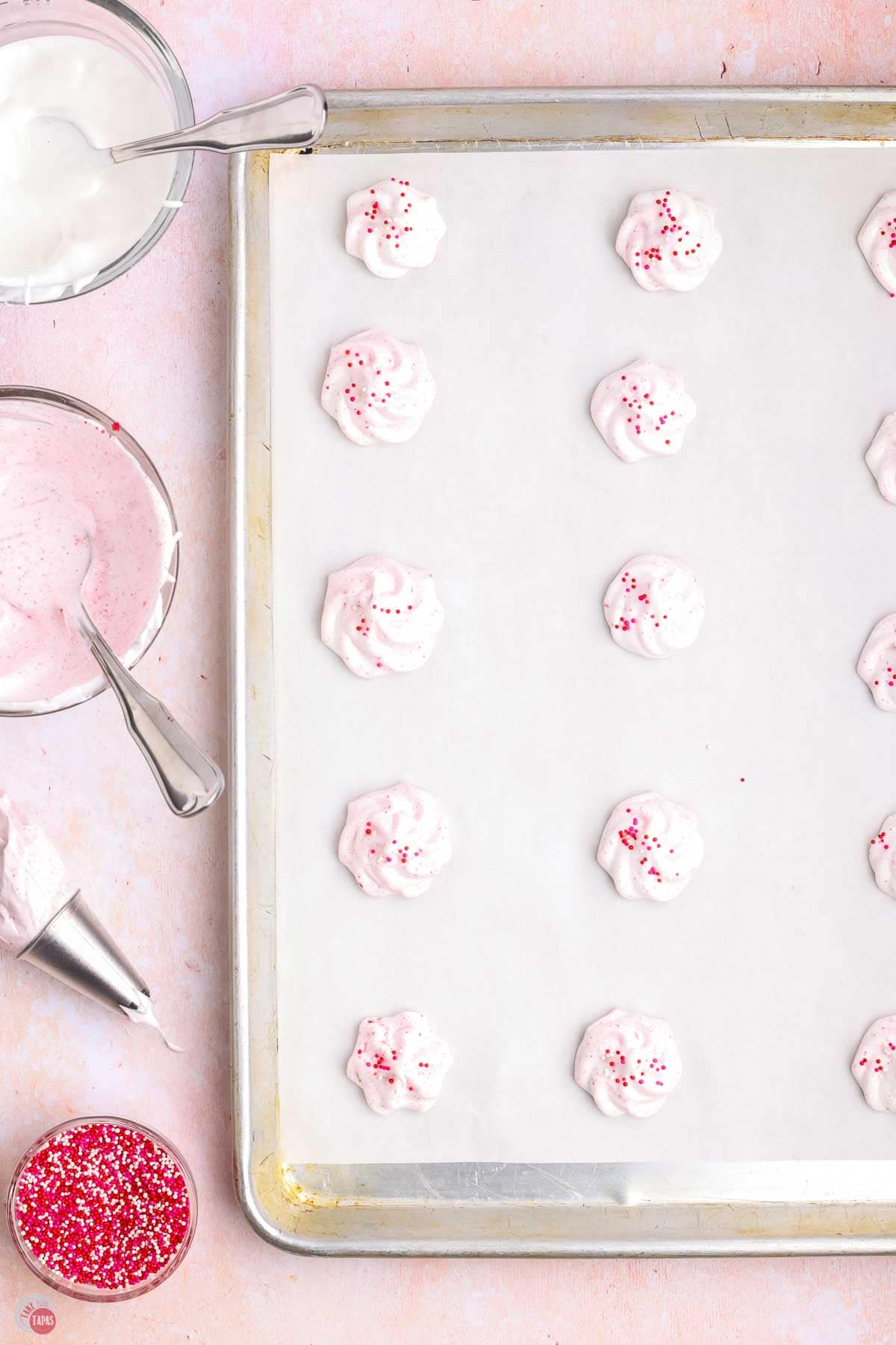pink meringues on a parchment lined baking sheet next to a bowl of sprinkles
