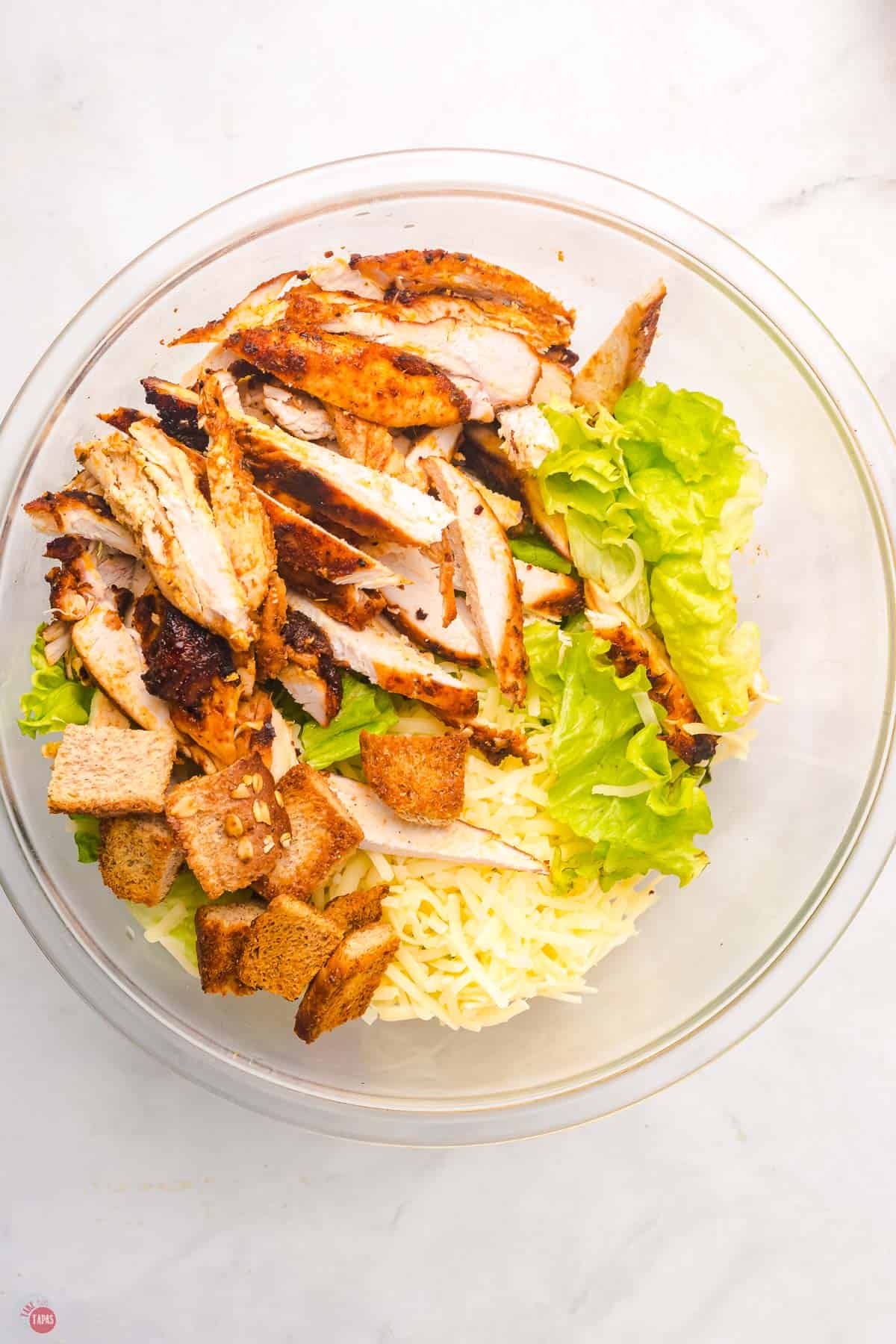 clear bowl full of lettuce, croutons, cheese, and sliced chicken