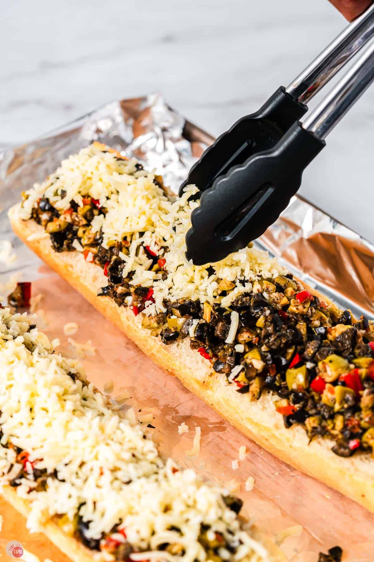 tongs placed shredded cheese on top of sliced bread with olives